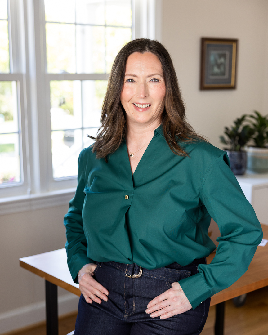 Smiling woman in a teal blouse stands with hands on hips in a bright room with a window above the table lighting.