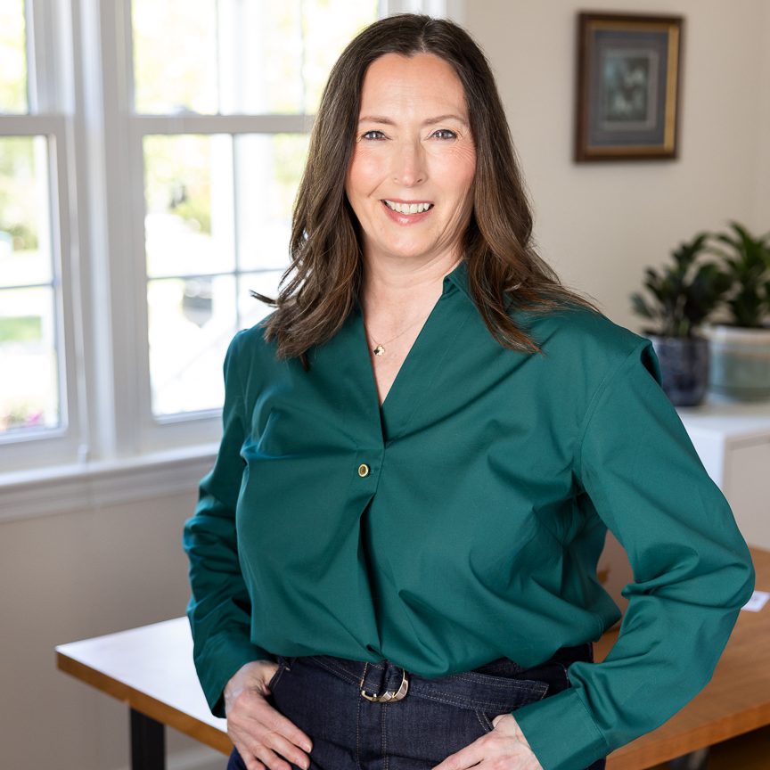 Smiling woman in a teal blouse stands with hands on hips in a bright room with a window above the table lighting.