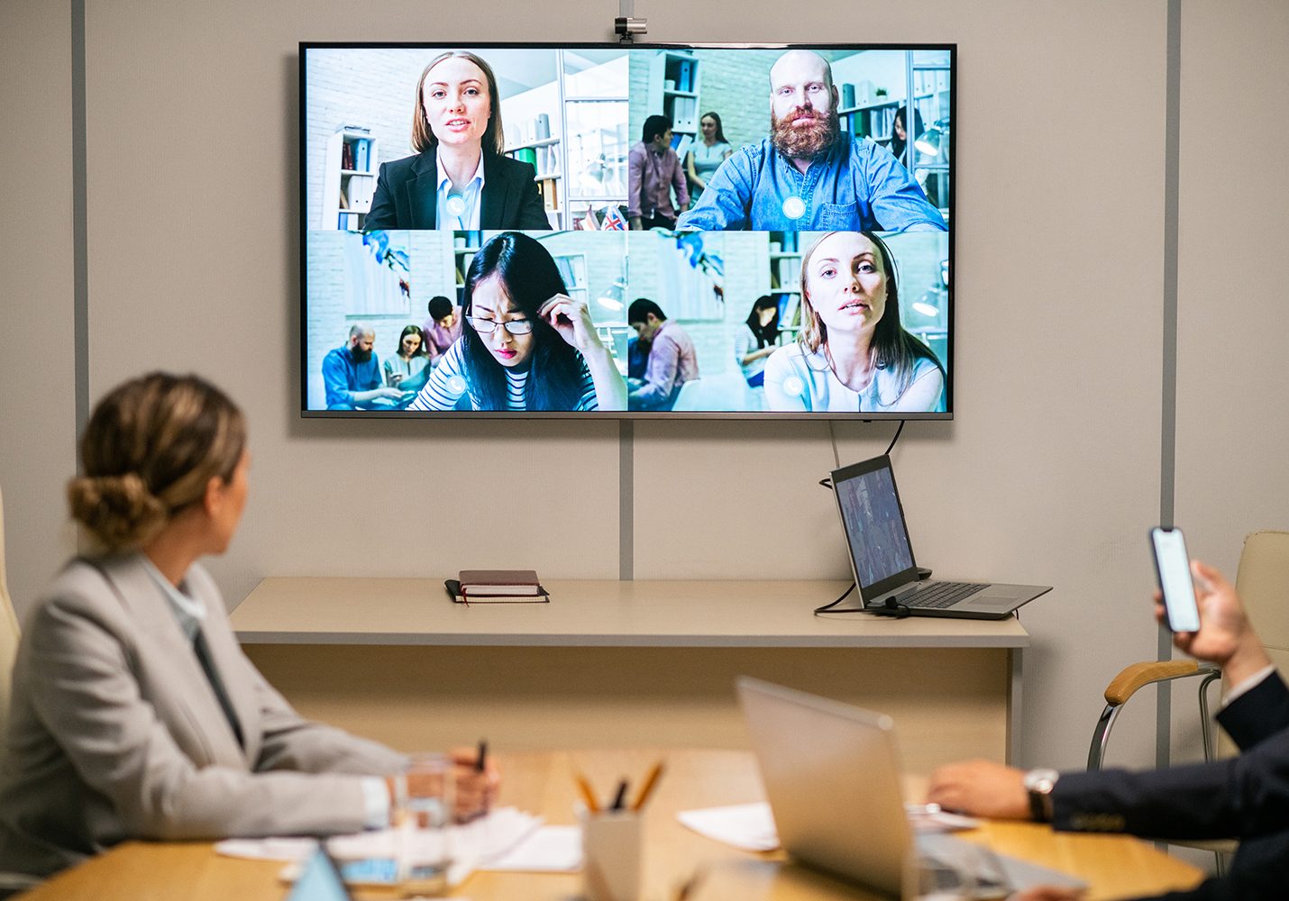 Meeting room with a wall screen showing a four-person video conference; attendees at the table, laptop and phone visible in foreground.